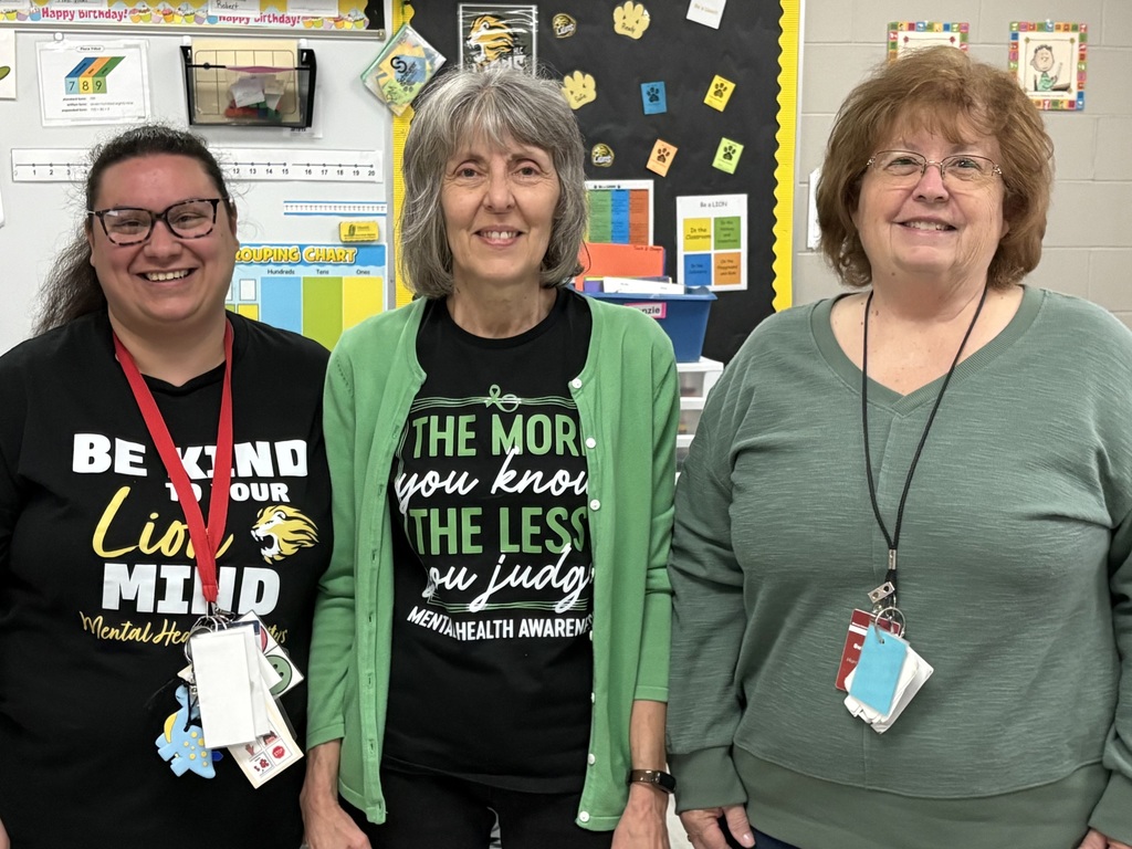 Three educators standing together indoors, smiling and wearing black shirts with mental health awareness slogans in green and white.