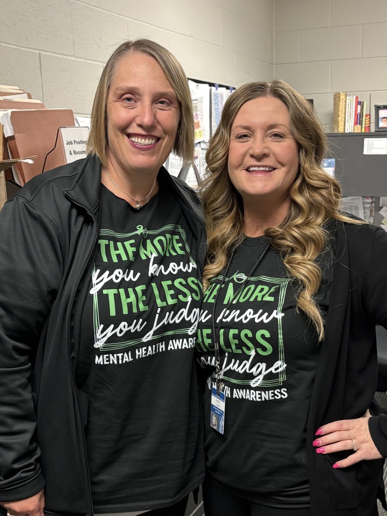 Two educators standing together indoors, smiling and wearing green and black mental health awareness shirts.