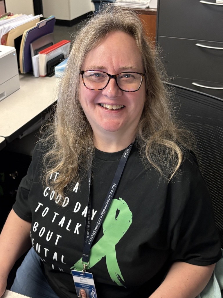 An educator smiling while seated at a desk, wearing a black shirt with a green ribbon and mental health awareness message.