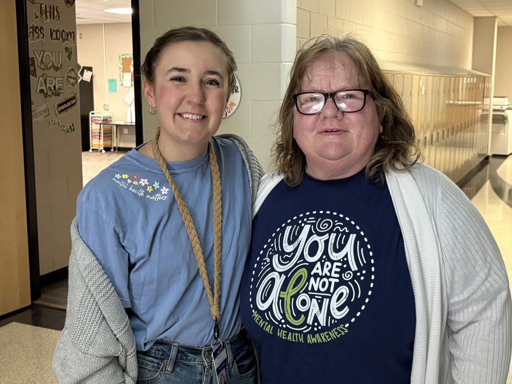 Two educators standing side by side against a hallway wall, smiling and wearing shirts that promote kindness and mental health awareness.