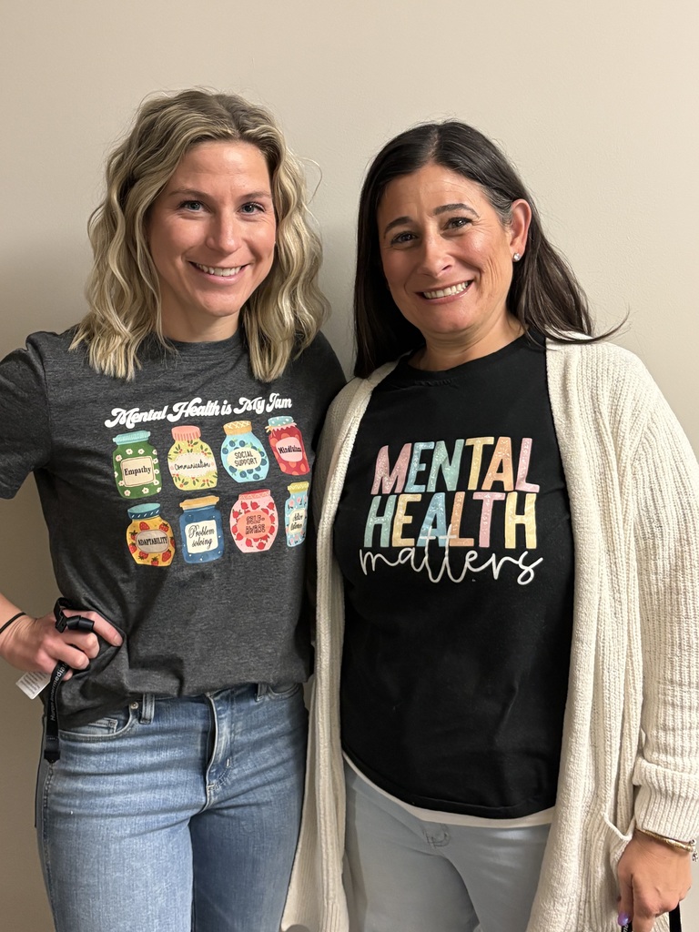 Two educators standing indoors, smiling and wearing shirts that promote kindness and mental health awareness.
