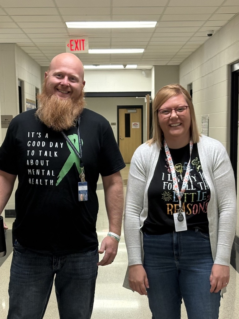 Two educators standing in a school hallway wearing black T‑shirts with green mental health awareness messages, smiling at the camera.
