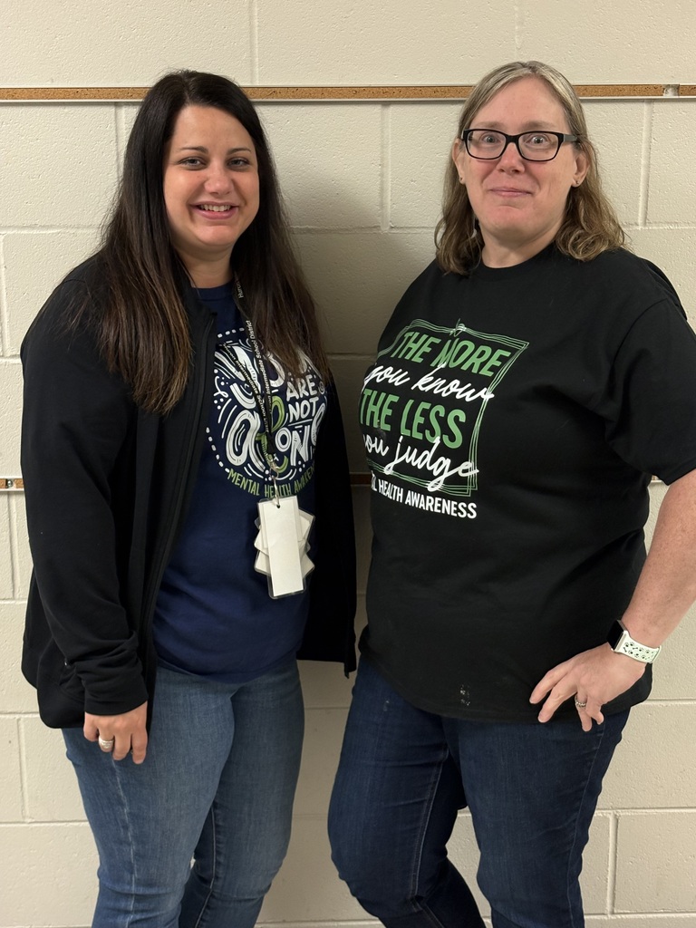 Two educators standing side by side in a hallway, smiling and wearing shirts that promote kindness and mental health awareness.