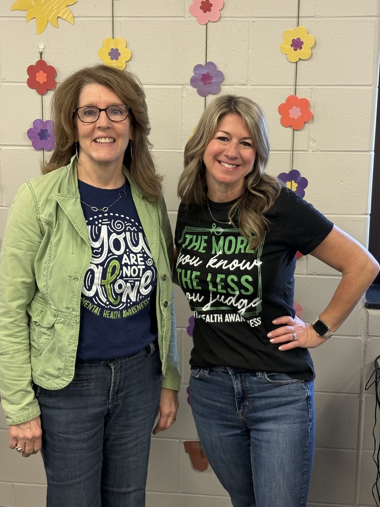 Two educators standing in front of a decorated wall with flower artwork, smiling and wearing mental health awareness shirts.