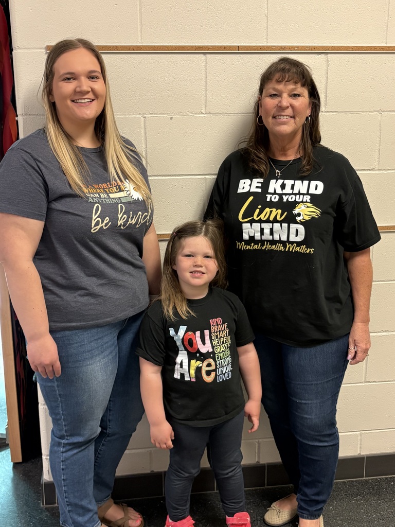 Two educators and a young child standing together against a wall, all wearing shirts with positive mental health messages.