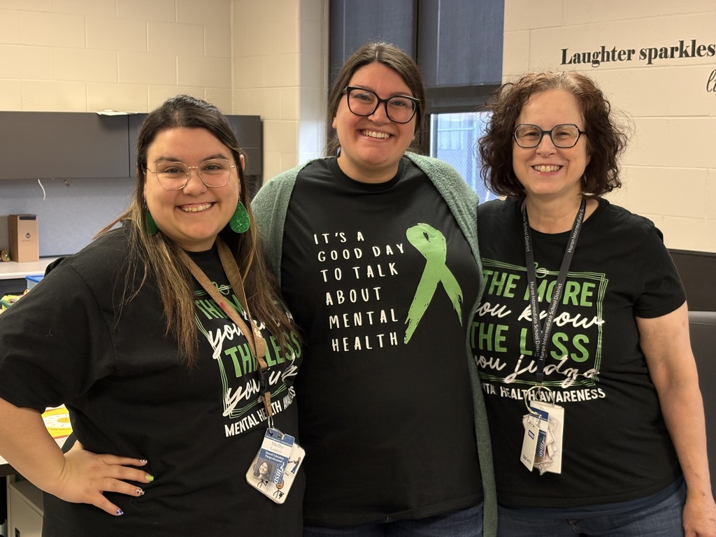 Three educators standing together indoors, smiling and wearing black shirts with mental health awareness slogans.