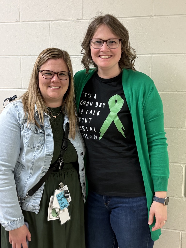 Two educators standing close together, smiling and wearing black shirts with green mental health awareness messages.