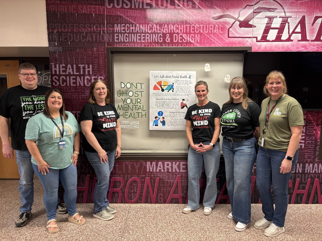 Seven HATC staff members stand side by side in a school hallway in front of a display about mental health. They are smiling and wearing green and black T‑shirts with messages promoting kindness and mental health awareness. Posters behind them include information about talking openly about mental health and ending stigma.