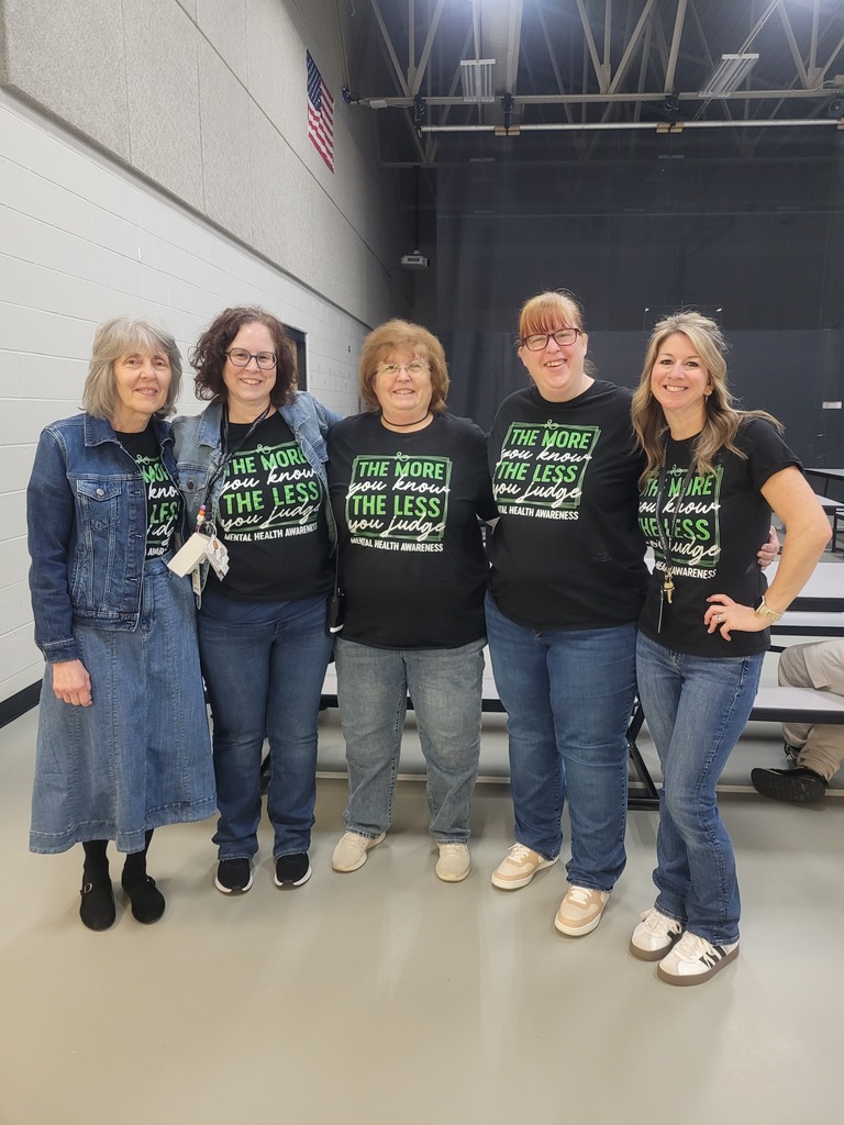 Five educators stand side by side indoors in a school multipurpose space. All are wearing black T‑shirts with green and white text that reads, “The more you know, the less you judge – Mental Health Awareness.” Several are wearing jeans. The background includes a light-colored wall, ceiling beams, and folded cafeteria tables.