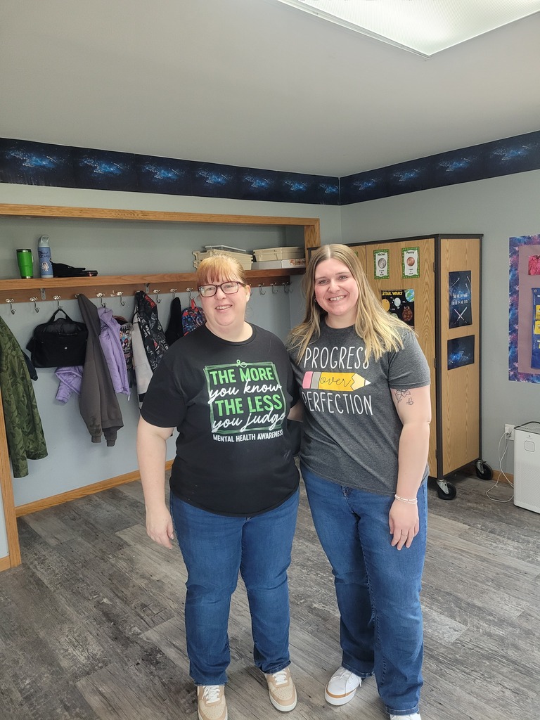 Two educators stand together inside a classroom. One is wearing a black “The more you know, the less you judge – Mental Health Awareness” T‑shirt, and the other is wearing a gray T‑shirt that reads “Progress over perfection” with a pencil graphic. The classroom background includes cubbies, hanging coats, posters, and educational materials.