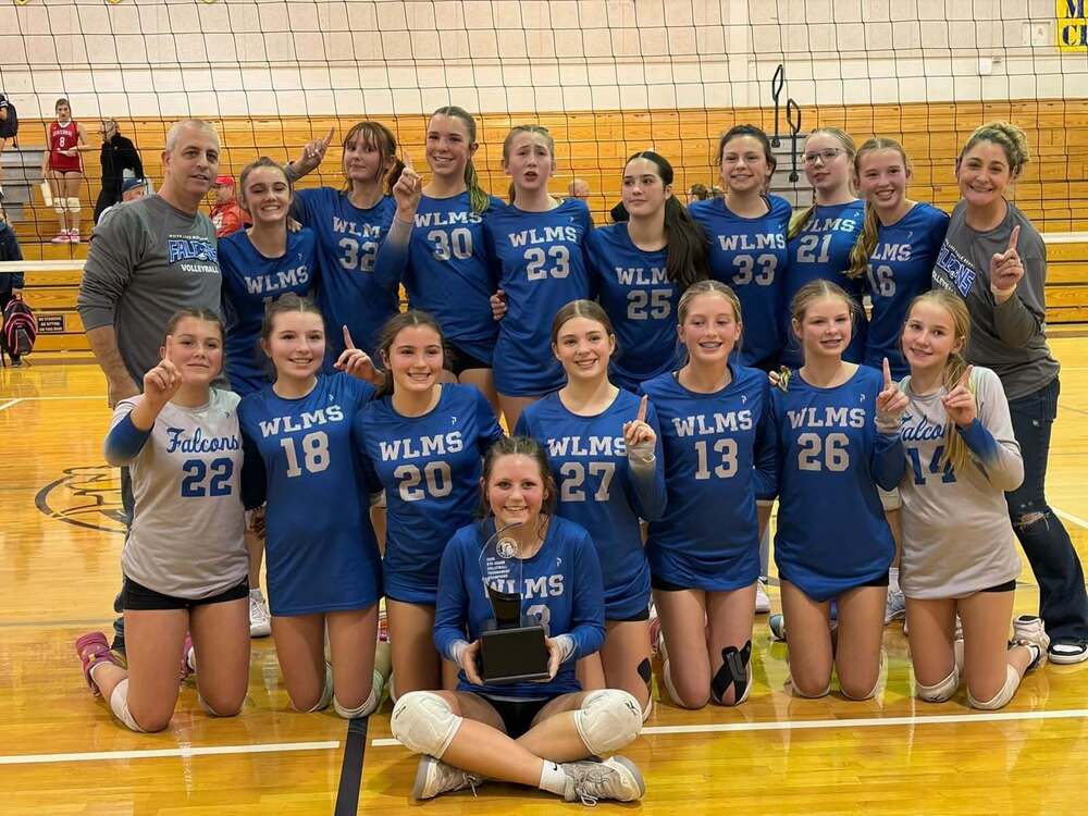 photo of girl volleyball team in blue shirts smiling and posing for a photo in front of a volleyball net