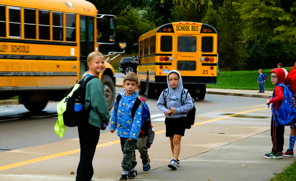 photo of girl leading young boys near the curb by school busses