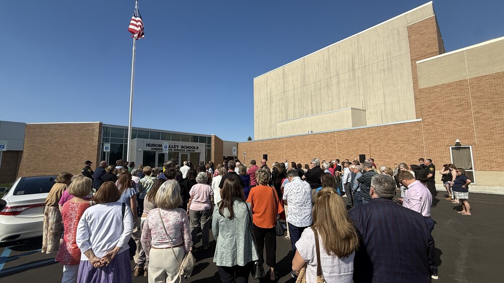 Dozens of people standing out front the new HVS admin center on a bright day listening to a speaker at a podium with the american flag overhead