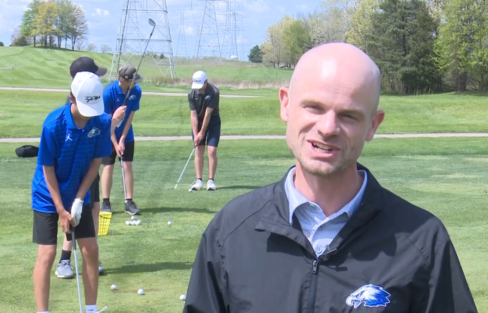 Photo of man looking at the camere while golfers practice behind him on the grass