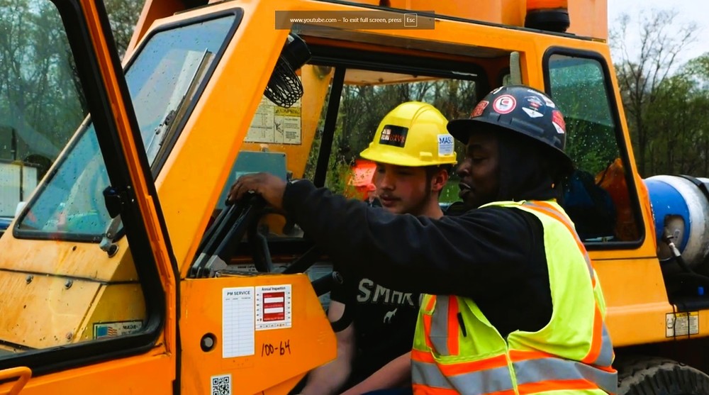 Student in hard hat being taught by adult how to run a large piece of machinary