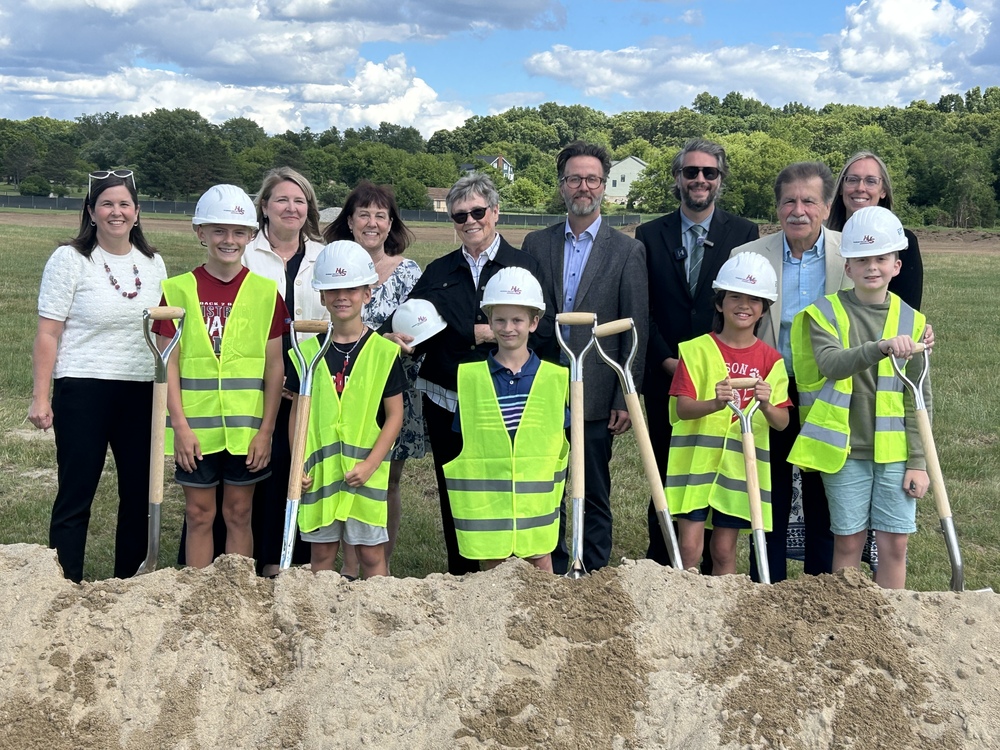 Group of students and adults standing behind dirt mound with shovels in hand