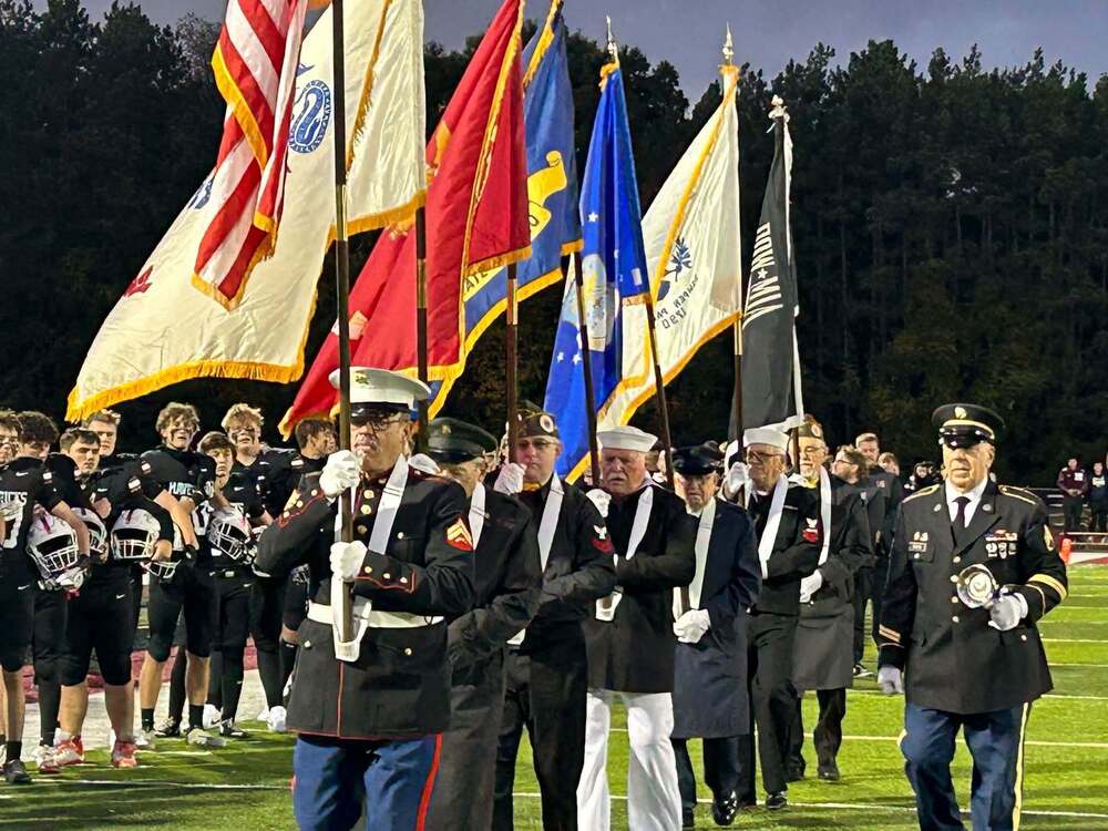 Several veterans acting as a color guard holding flags on a football field with players lined up on sidelines behind them
