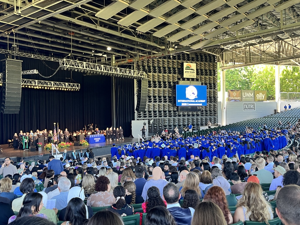 Photo of dozens of students in blue caps and gowns standing in the crowd at graduation day