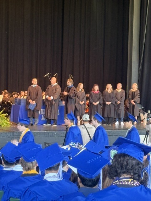 Educators on stage in grad gowns standing during graduation