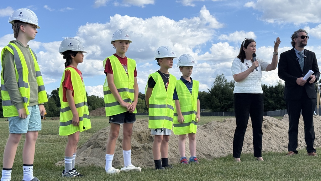 4 kids with safety vest  and hard hats on standing next to man and woman while the woman speaks into a microphone. 