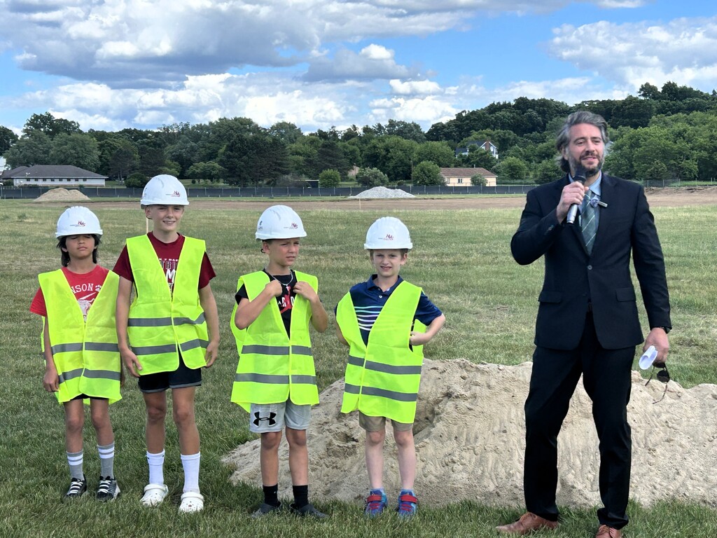 photo with 4 kids in safety vests next to a mound of dirt and a man in a suit speaking next to them 