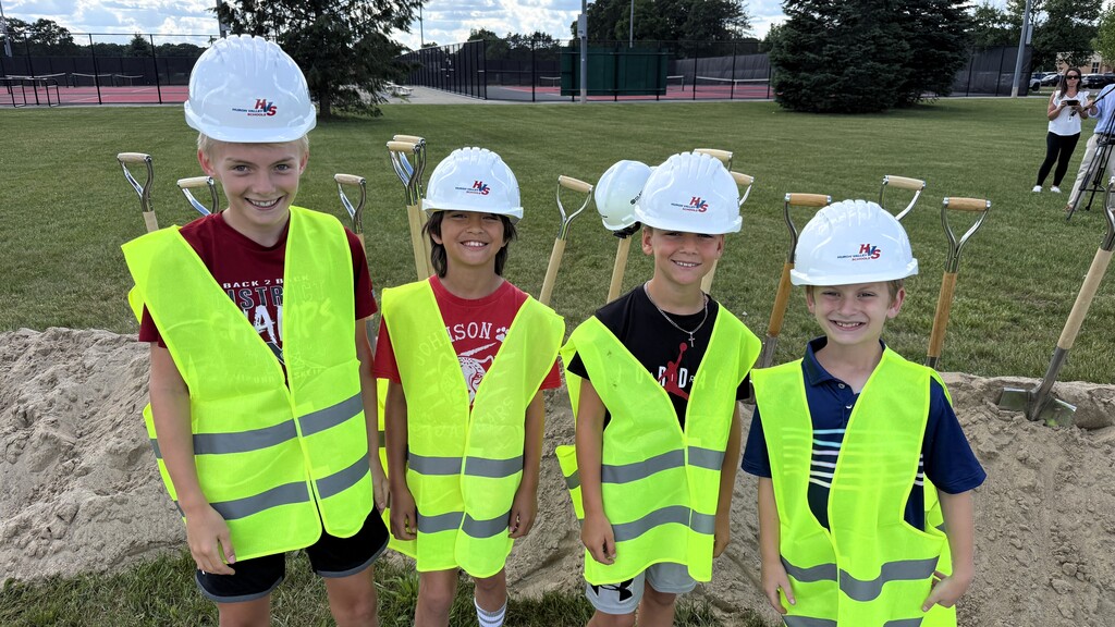 Group of 4 kids wearing safety vests, smiling with shovels behind them 
