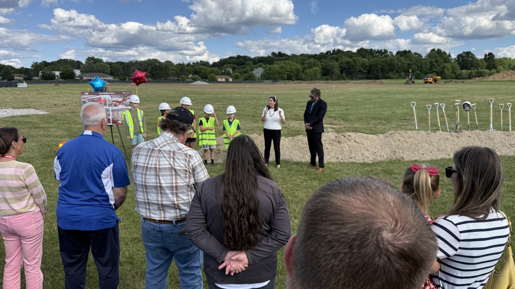Several people standing & listening to a man speak into a microphone near some kids in safety vests & a mound of dirt