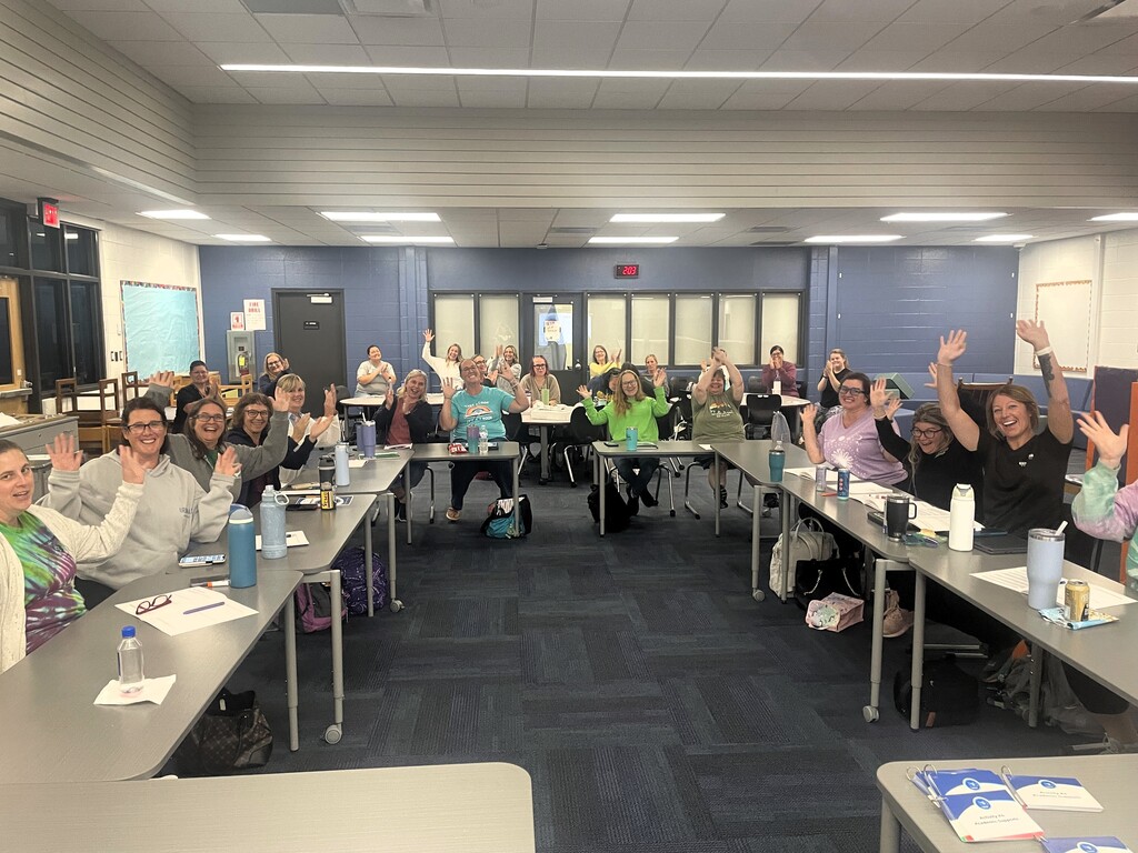 Photo of several women in a classroom setting sitting at a table & posing