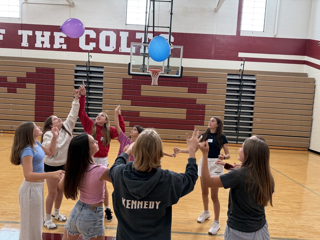 A circle of students inside a school gymnasium looking up and reaching their hands into the air toward two balloons floating overhead.