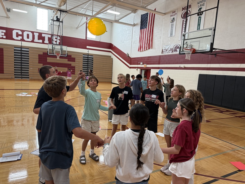 A group of students standing in a circle inside a school gymnasium, holding hands and looking up at a yellow balloon floating above them.