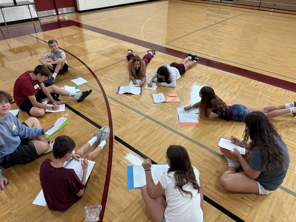 A group of students sitting and lying on the floor of a school gymnasium in a circle, working on worksheets in colorful folders.
