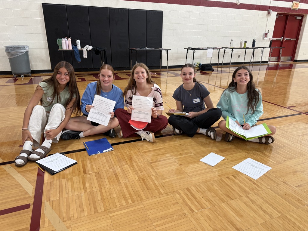 Five students sitting cross-legged in a row on the floor of a school gymnasium, smiling while holding papers and folders.