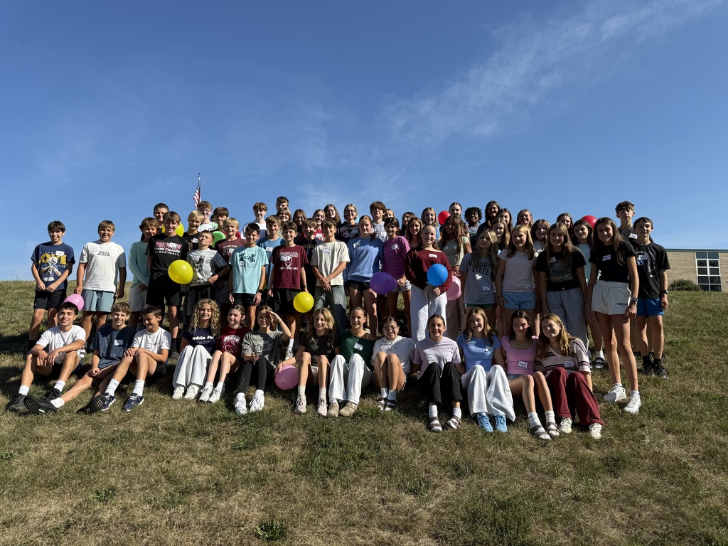 A large group of middle school students standing and sitting together on a grassy hill outdoors under a clear blue sky, with many holding colorful balloons.