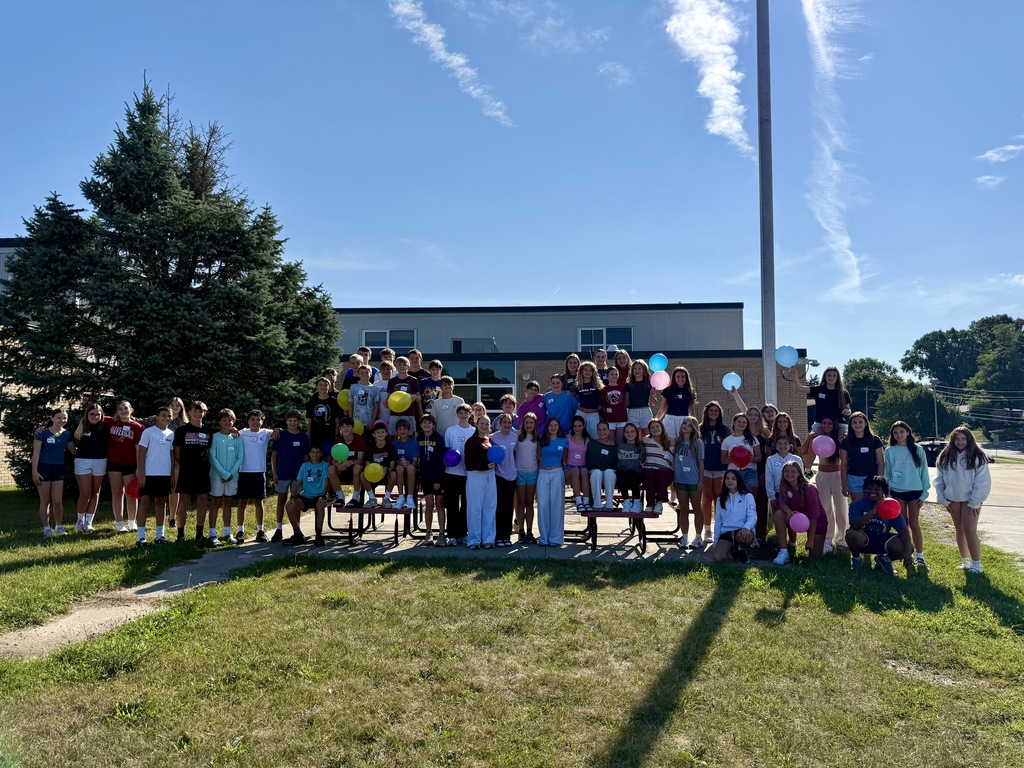 A large group of students posed outside in front of a school building on a sunny day, with several holding colorful balloons.
