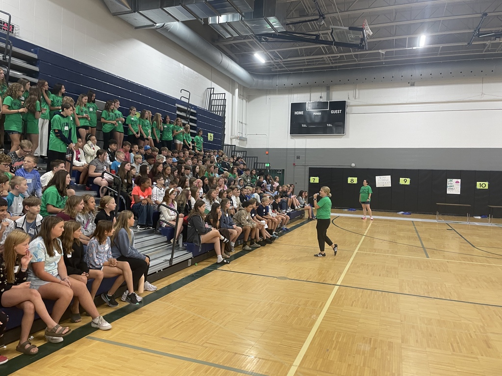 Adult speaking to dozens of kids/students sitting on bleachers in a gym