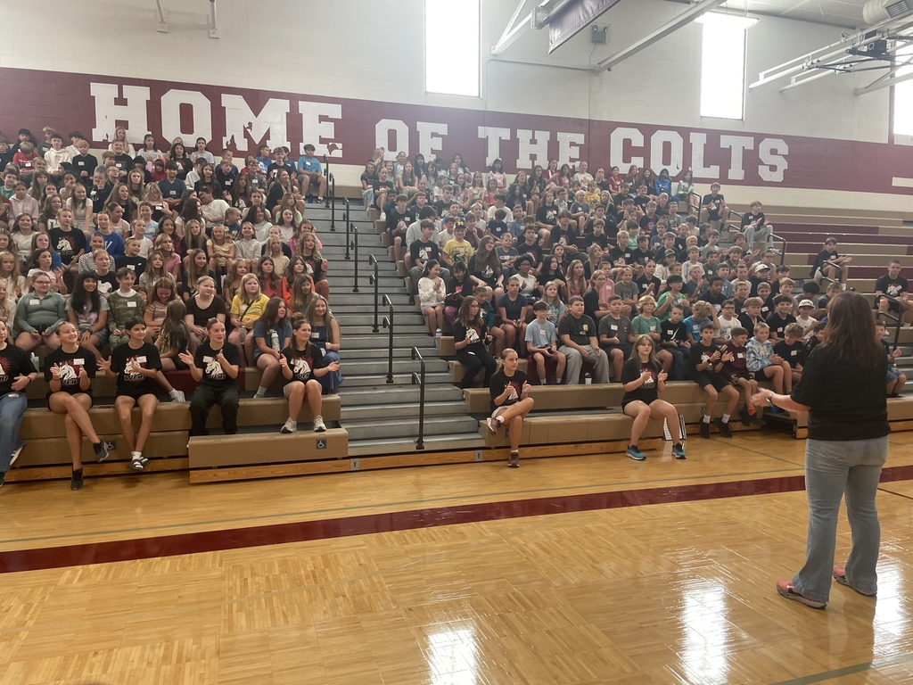 Adult speaking to dozens of kids/students sitting on bleachers in a gym