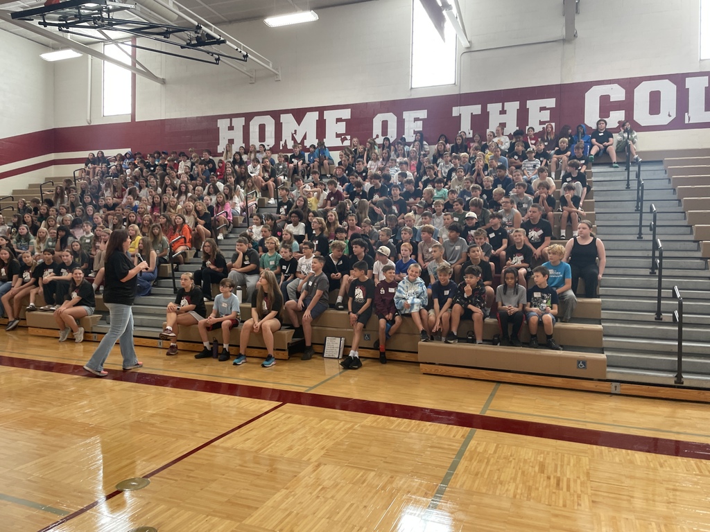 Adult speaking to dozens of kids/students sitting on bleachers in a gym