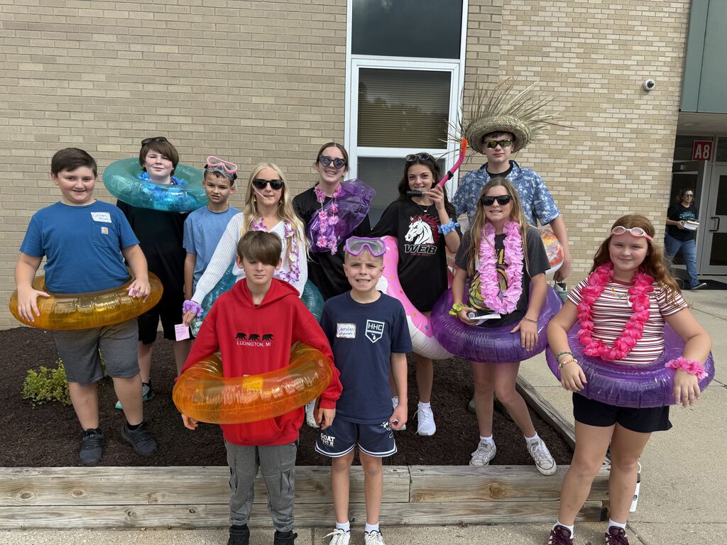 group of students posing outdoors dressed in beach gear, wearing colorful flower leis, swim goggles, and holding brightly colored inflatable pool tubes.