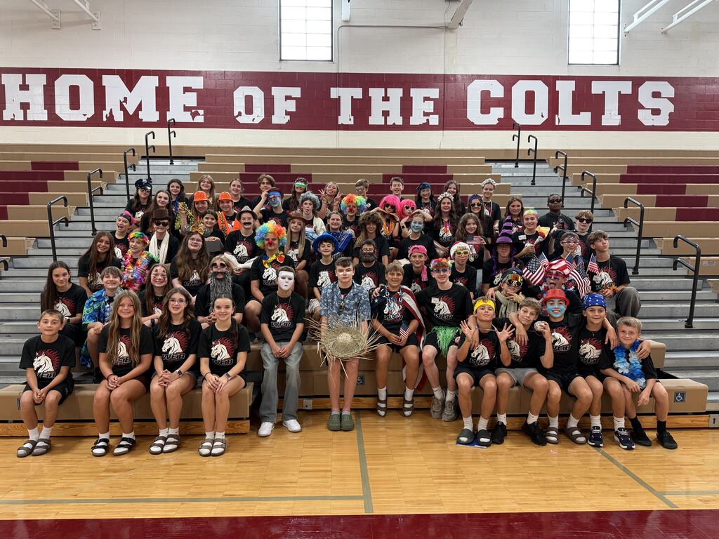 A large group of middle school students dressed in various themed costumes—including clown wigs, face paint, hats, and American flags—pose together on gym bleachers beneath a large wall sign reading "HOME OF THE COLTS."