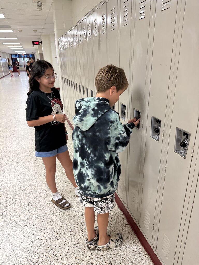 Two middle school students standing in a school hallway, with one student helping open a locker while the other looks on.