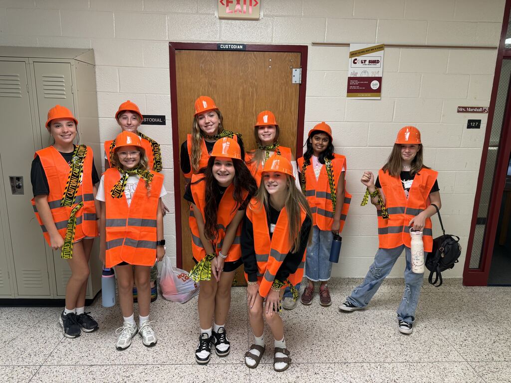 A group of nine middle school students posing in a hallway while wearing orange safety vests, orange hard hats, and yellow caution tape