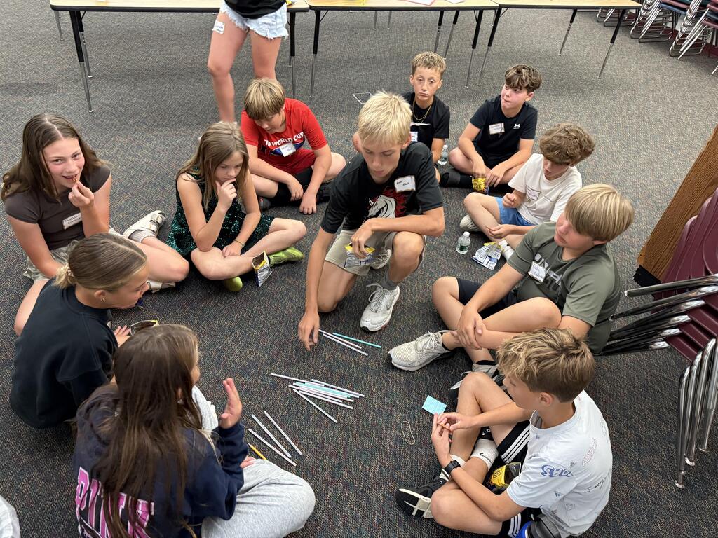 A group of students sitting in a circle on a carpeted floor during a classroom activity, working together with drinking straws and rubber bands spread out in the center.