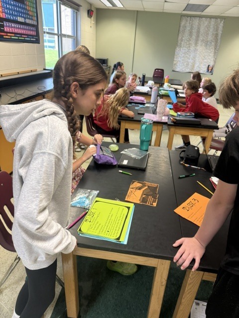 A student wearing a grey hoodie leans over a classroom table filled with colorful assignment sheets, school supplies, and laptops while working alongside classmates.