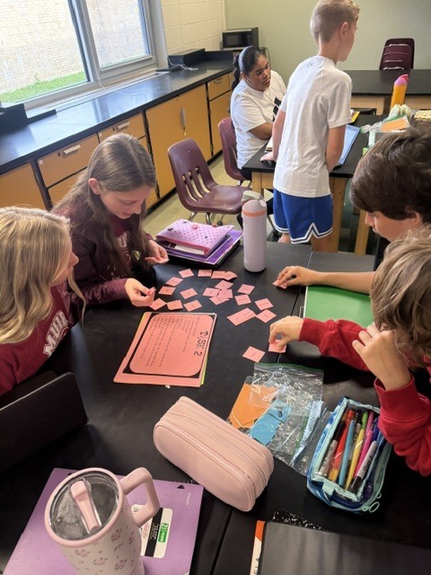 A top-down view of several students collaborating around a dark desk covered with pink task cards, pencil cases, water bottles, and learning materials.