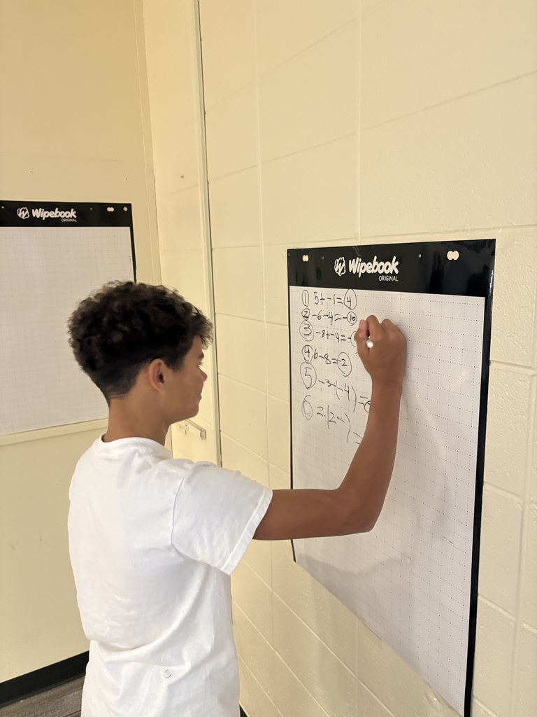 A student with short dark curly hair stands in profile, writing numbered math equations with a marker on a white reusable whiteboard grid attached to a classroom wall.