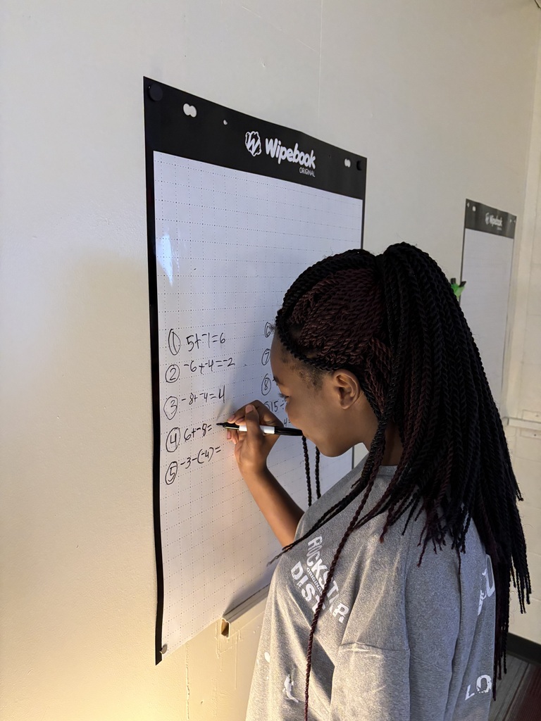 A student with long braided dark hair stands in profile, writing math problems with a dry-erase marker on a wall-mounted Wipebook poster.