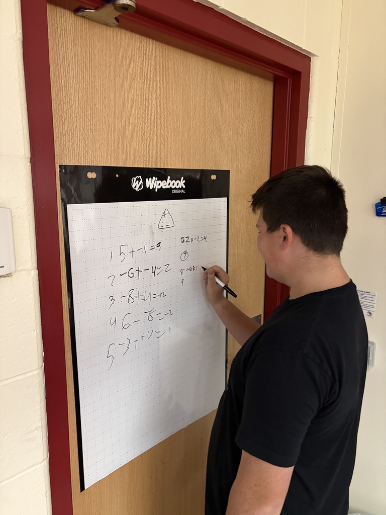 A student wearing a black t-shirt stands facing a classroom door, using a black marker to solve math equations on a Wipebook grid poster mounted to the door panel.