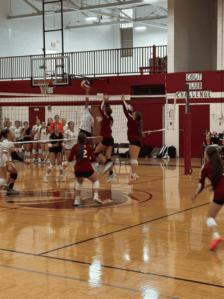 Action shot of a volleyball match in a gymnasium showing two players jumping high at the net to block a shot, with teammates positioned across the wooden court.