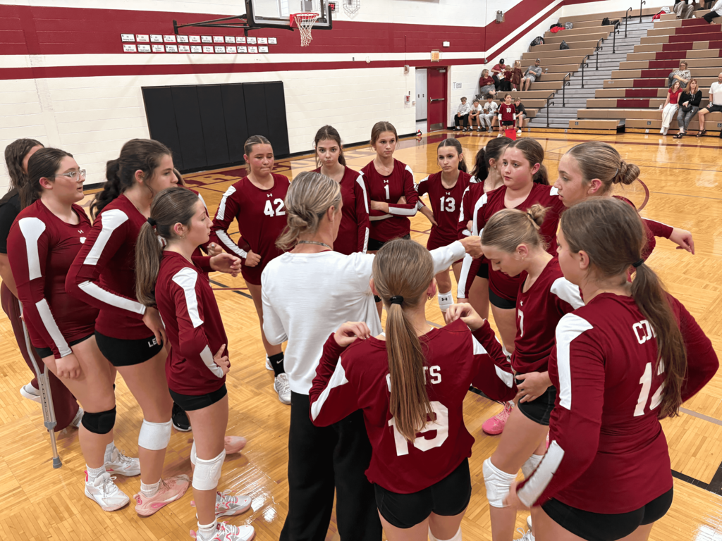 A volleyball team in maroon and white long-sleeve jerseys gathers in a tight huddle around their coach on a wooden gymnasium court.