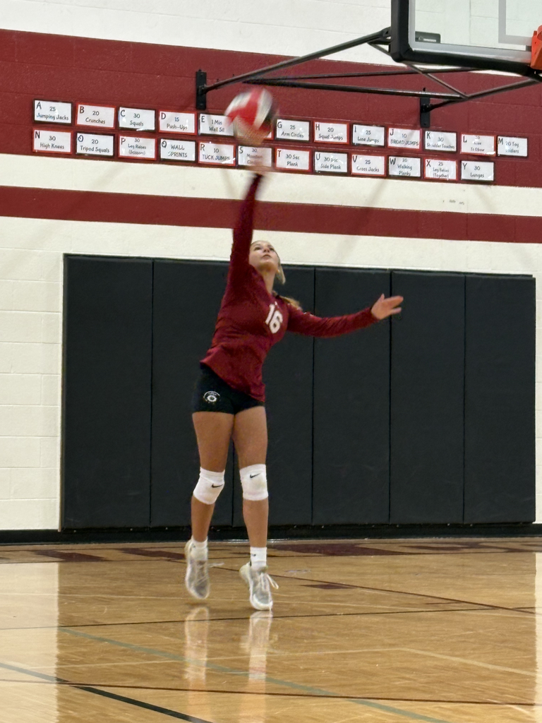 A volleyball player wearing jersey number 16 jumps mid-air on a basketball court, reaching high overhead with one hand to hit a volleyball.